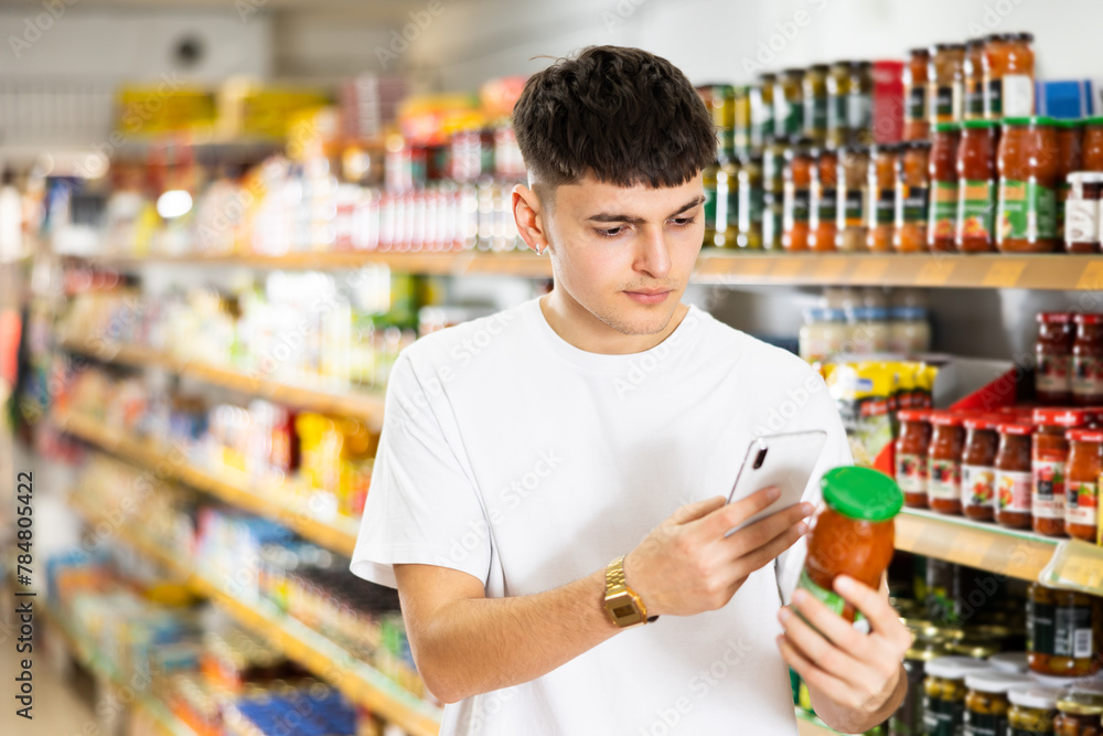 Focused interested young guy making purchases in supermarket, using smartphone to scan barcode on glass jar of canned food. Concept of modern technologies for shopping
