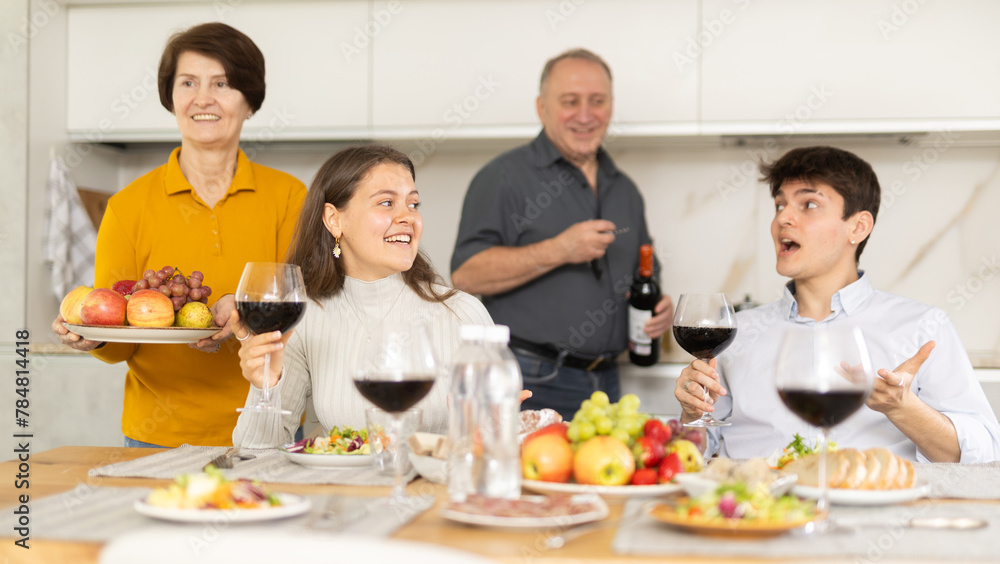Carefree young girl enjoying family dinner with husband and friendly aged parents-in-law, sharing stories and laughing over wine and appetizers in cozy kitchen