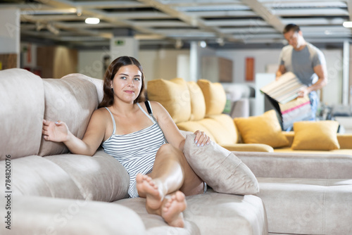 In spacious furniture store,girl is sitting on light settee and hugging pillow.It tests convenience and comfort of selected upholstered furniture