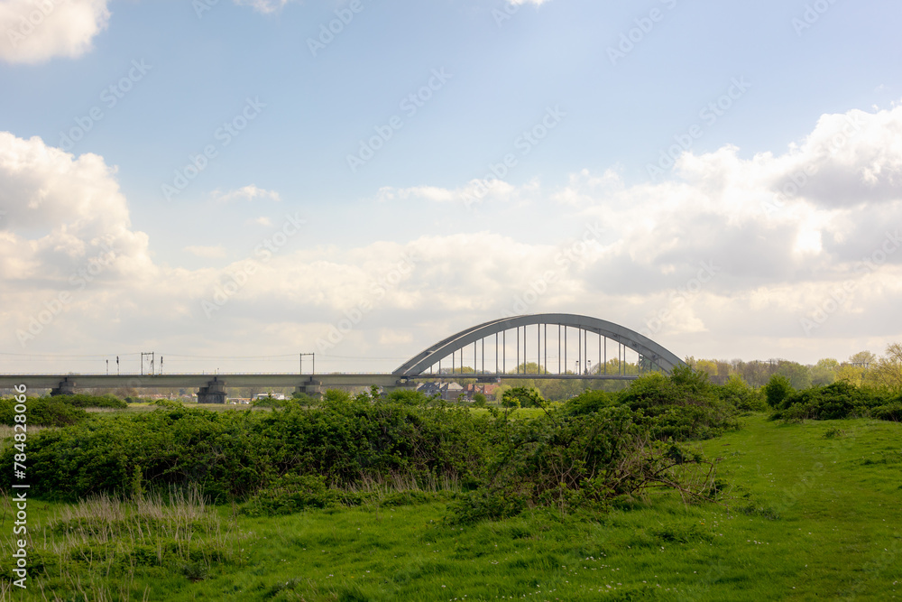 Obraz premium Spring landscape with green grass meadow along the Lek river, De Kuilenburgse spoorbrug (The Culemborg railway bridge) cross the river in a small village in the Dutch province of Utrecht, Netherlands.