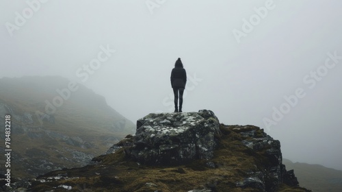 A person stands on a rocky outcrop back facing the camera as they bravely face the misty mountain pass leading to the rumored . .