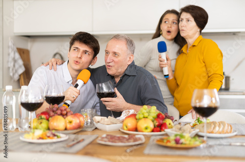 Cheerful lively multi-generational family  elderly parents and young adult children  singing into microphones  enjoying karaoke session at home dinner party with food and wine