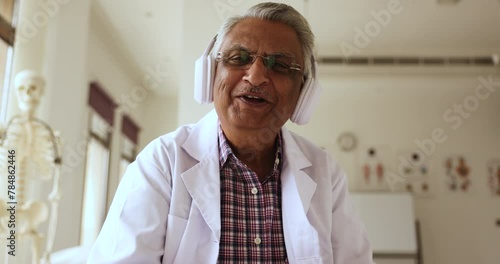 Happy elder Indian practitioner man in headphones talking on video call, to patient, speaking at camera, smiling, asking questions, giving medical recommendation, consultation photo