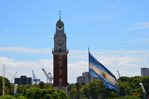 View of the Argentine flag, Torre Monumental or Torre de los Ingleses and Plaza General San Martin in Retiro with beautiful green trees, against a blue summer sky with some white clouds. Buenos Aires,