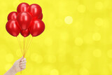 Female hand holding bunch of shiny red balloons on blurred yellow background. Empty space for text