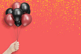 Female hand holds bunch of red and black balloons on red background with confetti. Empty space for text