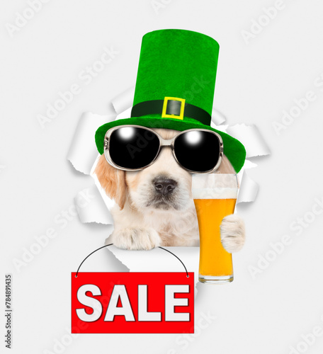 Saint Patrick's Day concept. Cute Golden retriever puppy wearing green hat of the leprechaun looks through the hole in white paper, holds mug of light beer and shows signboard with labeled "sale"