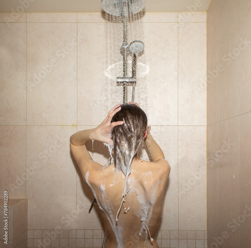 Back view of anonymous naked female standing in shower cabin with brick tiled wall. She is lathering shampoo on head while washing brown hair. UGC