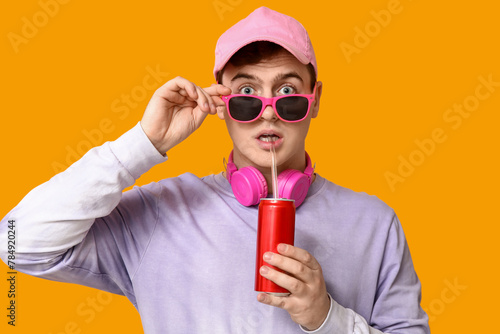 Canvas Print Shocked young man in sunglasses drinking soda on yellow background