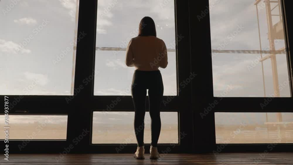 Low angle approach toward female stand near wide studio apartment window Low angle approach toward female stand near wide studio apartment window