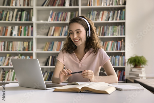 Happy girl in headphones engaged in on-line class, listening to tutor, make homework, doing schoolwork sit at desk with laptop, studying in college library on bookshelves background. Education, tech