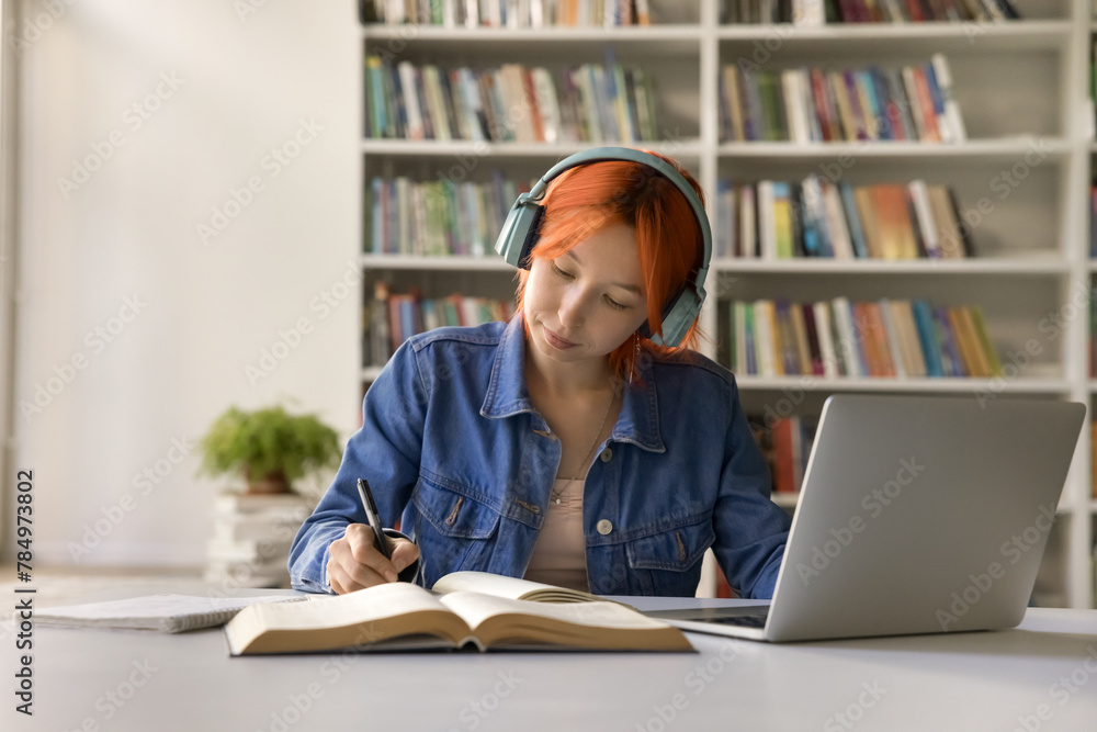 Focused red haired 17s student girl doing schoolwork, listen audio course improving foreign language skills, e-learning on-line, makes notes, jotting information sitting at desk in high-school library Focused red haired 17s student girl doing schoolwork, listen audio course improving foreign language skills, e-learning on-line, makes notes, jotting information sitting at desk in high-school library