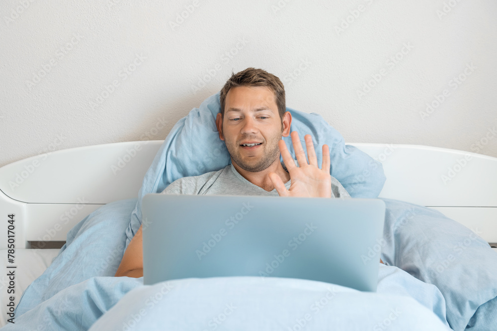 Young cheerful man communicate by video call with collegues shows greeting gesture while lying in the bed and working on a laptop at home. Happy Caucasian male freelancer have video conference Young cheerful man communicate by video call with collegues shows greeting gesture while lying in the bed and working on a laptop at home. Happy Caucasian male freelancer have video conference