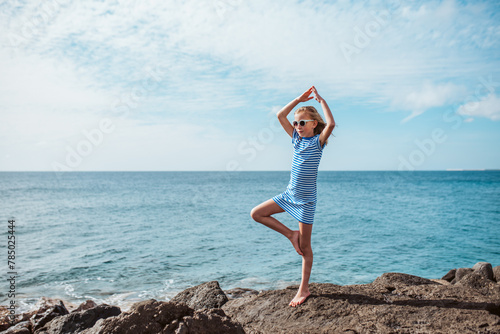Young girl standing on one leg on rock. Blonde girl in dress enjoying vacation in Canary Islands. Concept of beach summer vacation with kids.