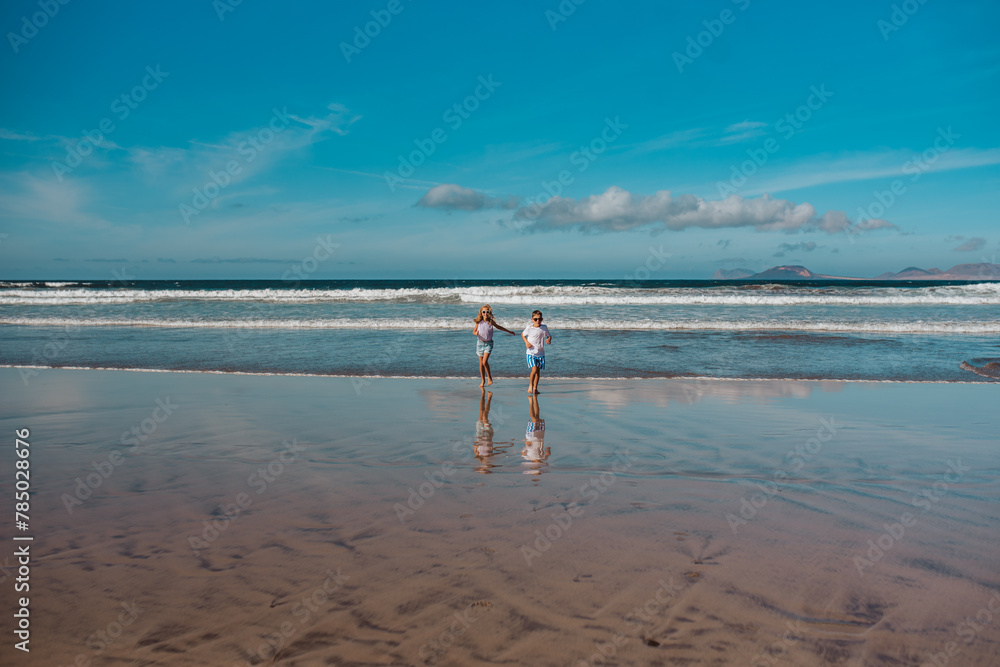 Siblings playing on beach, running, skipping in water. Smilling girl and boy on sandy beach of Canary islands. Concept of family beach summer vacation with kids.