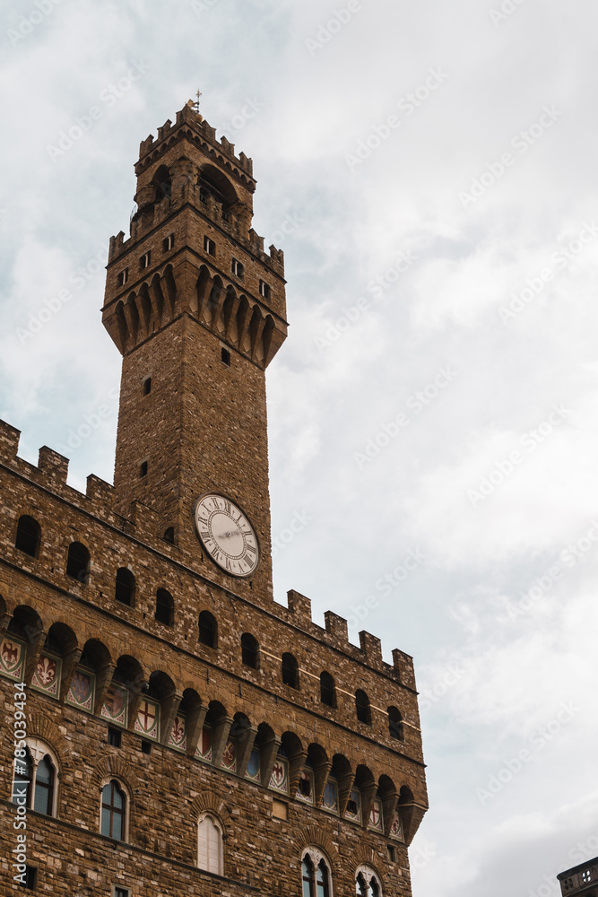 Fototapeta premium Palazzo Vecchio's imposing façade, adorned with medieval shields, from Piazza della Signoria. Torre di Arnolfo, with a clock and crowned with battlements, stands tall against a grey cloudy sky.