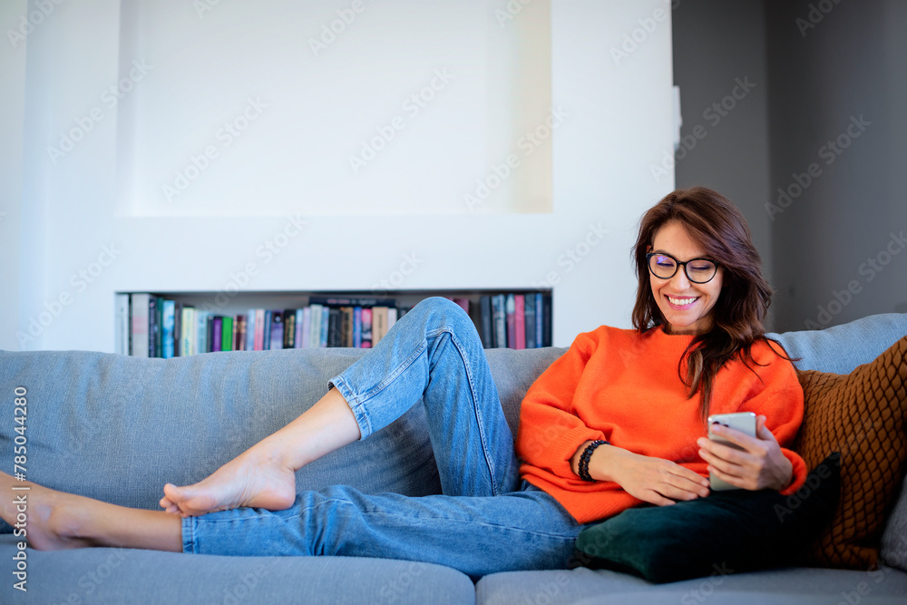 Fototapeta premium Full length of a middle-aged woman using her smartphone and lying on the sofa at home.