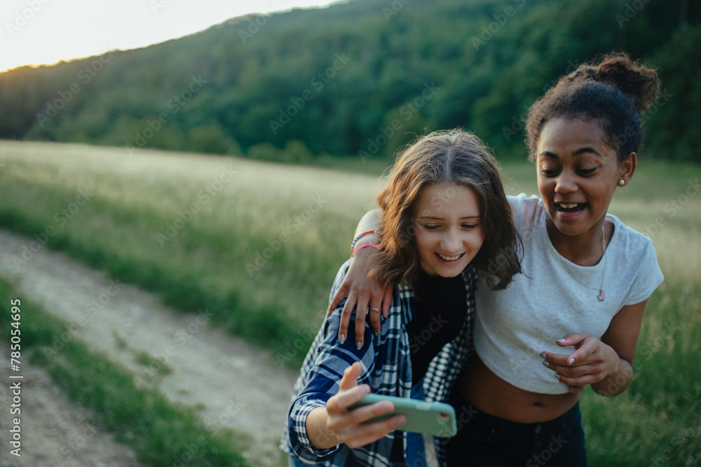 Young teenager girl best friends spending time in nature, during sunset. Girls on walk, taking selfie with smartphone. - obrazy, fototapety, plakaty 