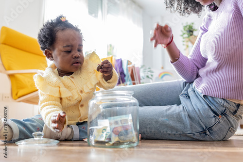 Young mother sitting near daughter playing with coins by jar of currency at home photo
