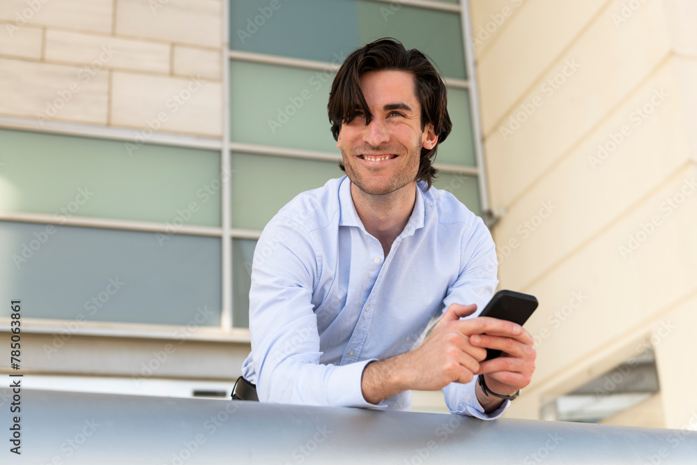 Smiling thoughtful businessman with smart phone leaning on railing