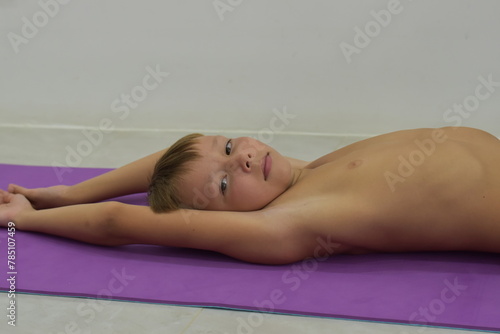 A blond-haired boy lies on a yoga mat with his arms outstretched.