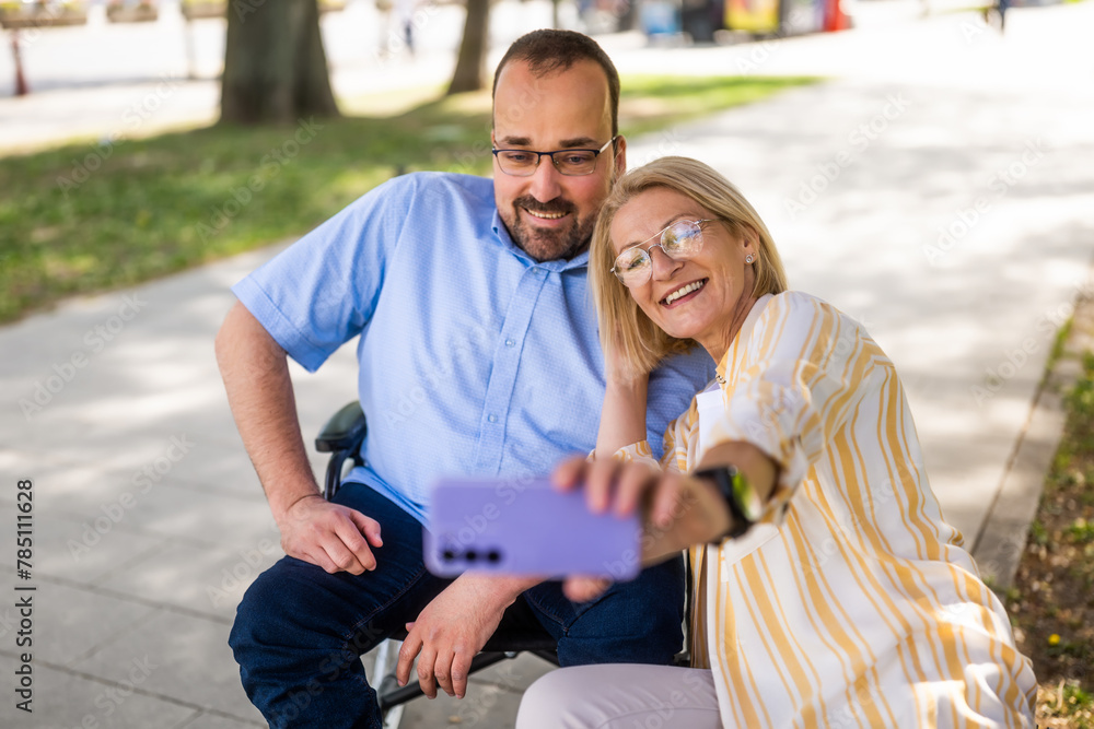 Man in wheelchair is spending time with his mother in park. They are taking selfie. Man in wheelchair is spending time with his mother in park. They are taking selfie.