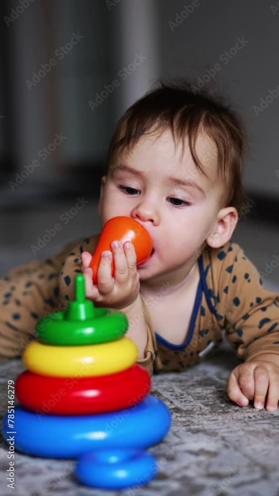 Baby boy in brown romper lies on the carpet indoors. Adorable one-year old child playing with a toy pyramid. Blurred backdrop. Vertical video Baby boy in brown romper lies on the carpet indoors. Adorable one-year old child playing with a toy pyramid. Blurred backdrop. Vertical video
