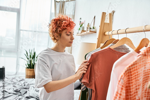 attractive red haired extravagant person picking up stylish clothes near rack at home, leisure time