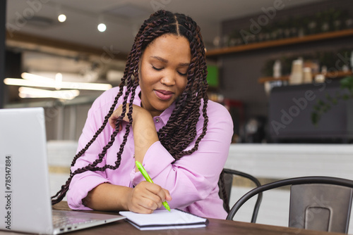 Biracial woman wearing pink shirt writing in notebook in a modern business office