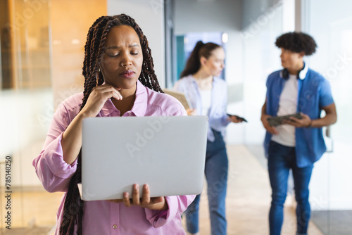 Biracial woman holding laptop, thinking, colleagues walking behind in a modern business office