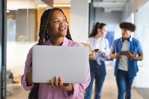 A young biracial woman holding a laptop, colleagues chatting behind in a modern business office