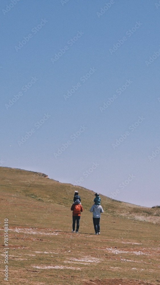 Vertical back view of two males holding little girls on their shoulders, walking on a flat terrain