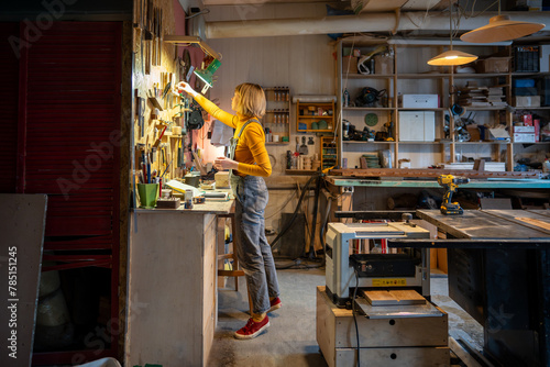 Focused on workflow in carpentry shop craftswoman carpenter in denim picks up tools to produce handmade wooden furniture. Small artisan business. Concentrated female cabinetmaker busy joinery workshop