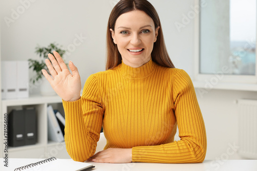 Woman waving hello during video chat at table in office, view from web camera
