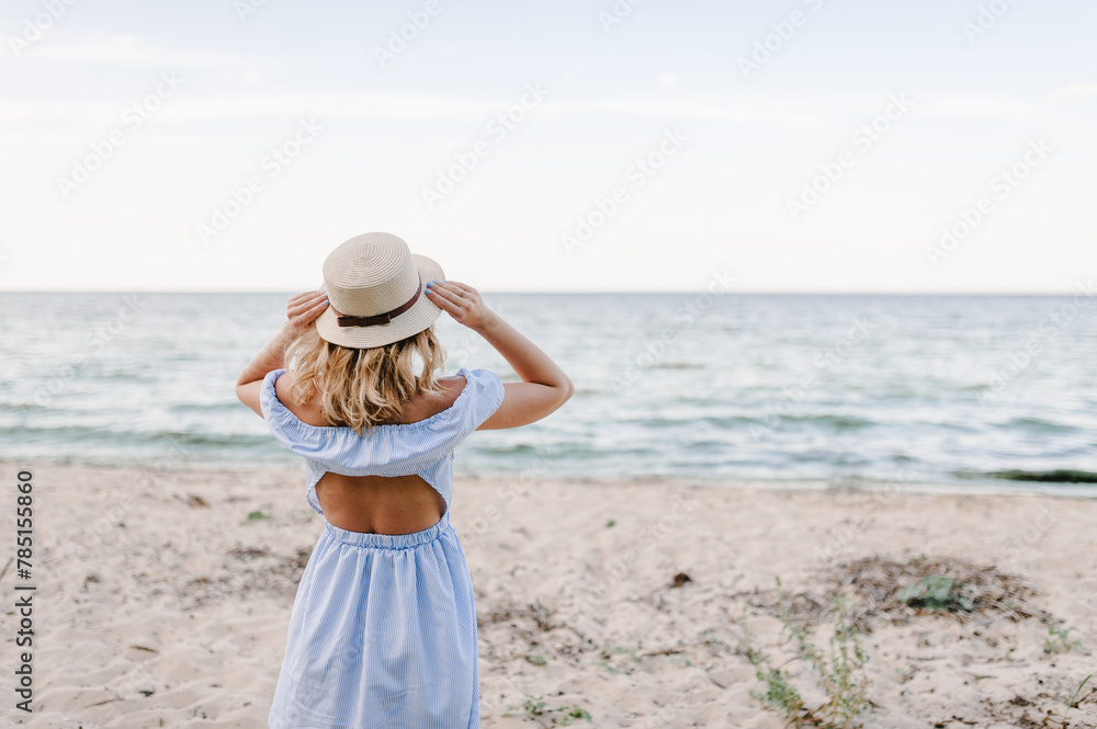 Portrait of stylish female on sand. Beautiful girl standing on beach ocean and enjoying sunny summer happy day on vacation. Back view. Young attractive blond woman in straw hat joyfully walks near sea Portrait of stylish female on sand. Beautiful girl standing on beach ocean and enjoying sunny summer happy day on vacation. Back view. Young attractive blond woman in straw hat joyfully walks near sea