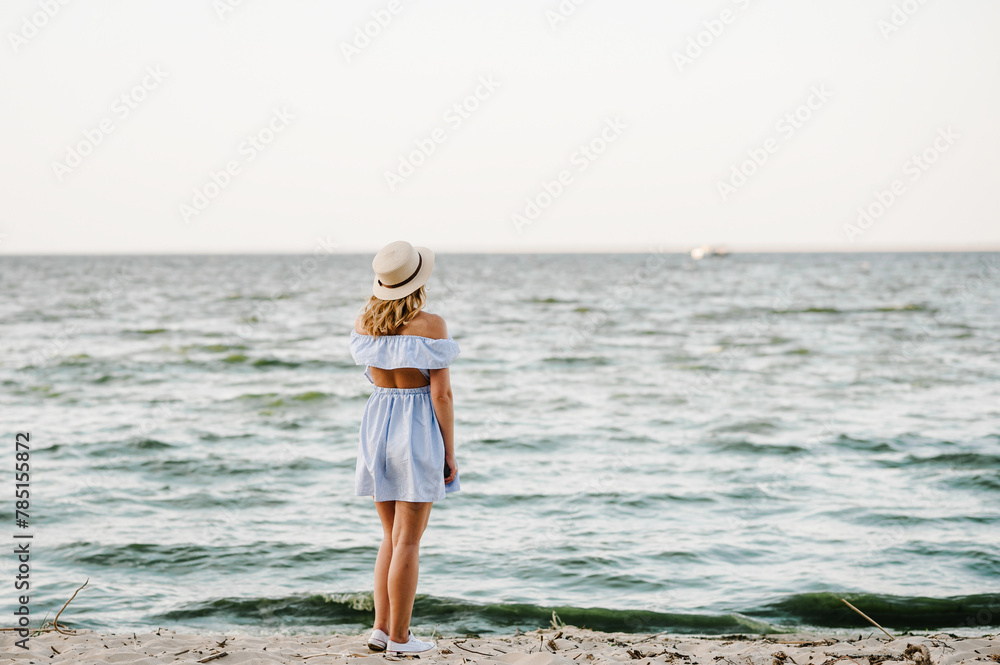 Young attractive blond woman in straw hat joyfully walks near sea. Portrait of stylish female on sand. Beautiful girl standing on beach ocean and enjoying sunny summer happy day on vacation. Back view