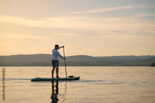 A man is standing and paddling on a paddleboard on the water at sunset.