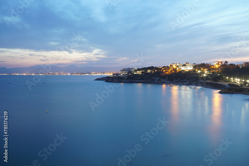 Tranquil Coastal Twilight Scene With Calm Waters and Illuminated Shoreline. A serene twilight overtakes the coast as the calm sea mirrors a pastel sky. Lights from the shoreline buildings twinkle