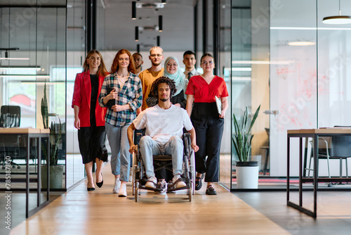 A diverse group of young business people walking a corridor in the glass-enclosed office of a modern startup, including a person in a wheelchair and a woman wearing a hijab