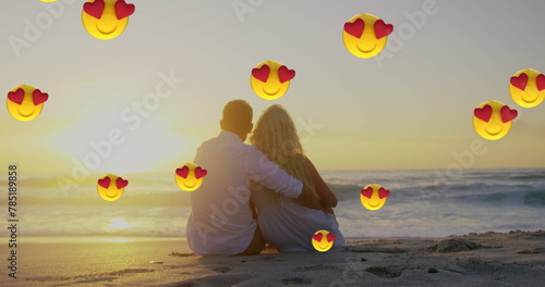 Image of multiple heart eyes face emoji floating against couple in love embracing sitting on beach