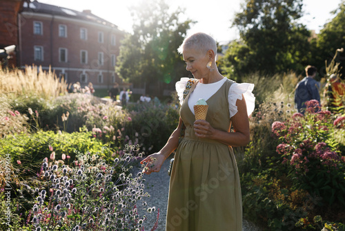 Outdoor side view image of beautiful romantic senior female with stylish look, wearing fashionable dress, walking in park, touching flowers and plants while eating tasty ice cream in waffle cone