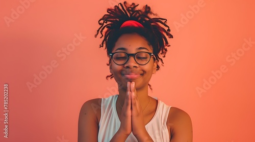 A happy millennial woman with glasses, posing with folded hands, on a solid peach background, radiating positivity