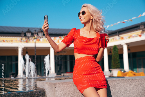 Young beautiful smiling hipster woman in trendy summer clothes. Carefree model posing in the street. Holding and drinking fresh vegetable cocktail smoothie drink in plastic cup with straw. Sunglasses