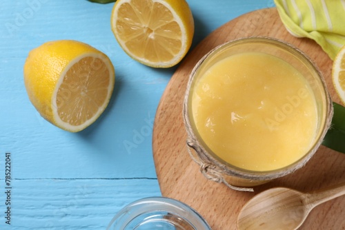Delicious lemon curd in glass jar, fresh citrus fruit and spoon on light blue wooden table, top view