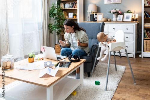 Young mother, business woman having video call on laptop from home while taking care of her baby sitting in tall baby chair. Female business owner on maternity leave must work online from home. © Srdjan