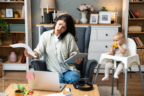 Young mother, business woman having cellphone conversation from home while taking care of her baby sitting in tall baby chair. Female business owner on maternity leave must work online from home.