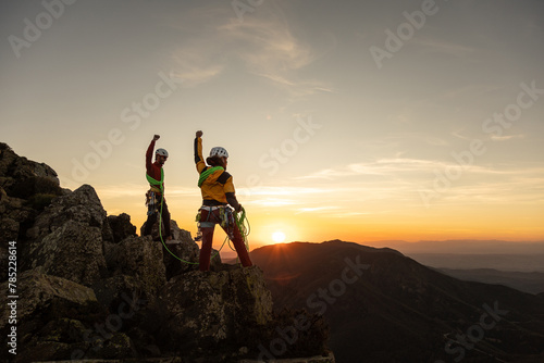 Two people are standing on a mountain top, one of them is holding a fist up in the air. The sun is setting in the background, creating a beautiful and serene atmosphere