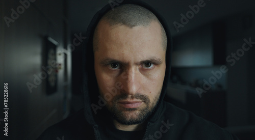 Front view portrait of a brutal man looking intently at the camera from under a black hood. Dark background. People and emotions.