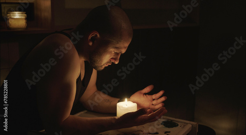 Side view of an serious sad adult caucasian man looking to the burning candle sitting in the dark room. Electricity power failure