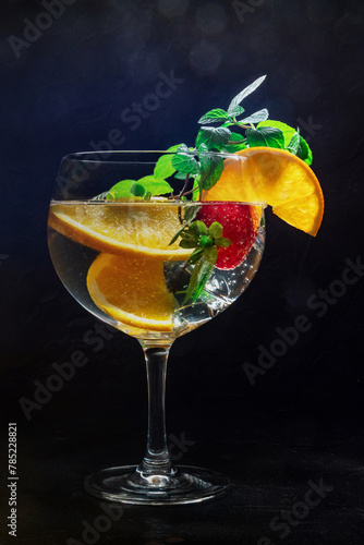 Fancy cocktail with fresh fruit. Gin and tonic drink with ice at a party, on a black background. Alcohol with orange, mint, and strawberry, toned image